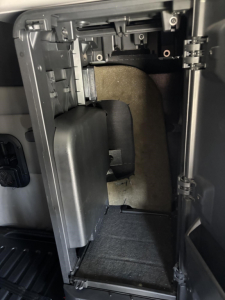 Cab interior area showing HVAC or storage compartment under dashboard with plastic trim and wiring harness; empty small cavity with dark carpeting.