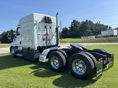 Freightliner Cascadia 126 sleeper tractor from left-rear angle showing dual rear axles, white tractor cab, external air tanks, exhaust stack, and fifth-wheel with mudflaps.