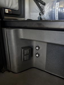 Cab area of a Freightliner Cascadia showing a dual USB/aux panel and two round buttons near a storage compartment.