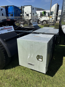 Aluminum toolbox mounted on side of Freightliner Cascadia truck chassis with fuel/air tanks and parked trailers in background.