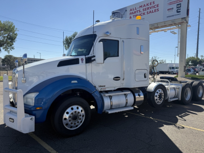 Kenworth T880 truck in white and blue, front view with large bumper and dual exhaust, cab with sleeper, five wheels visible on rear, billboard in background.