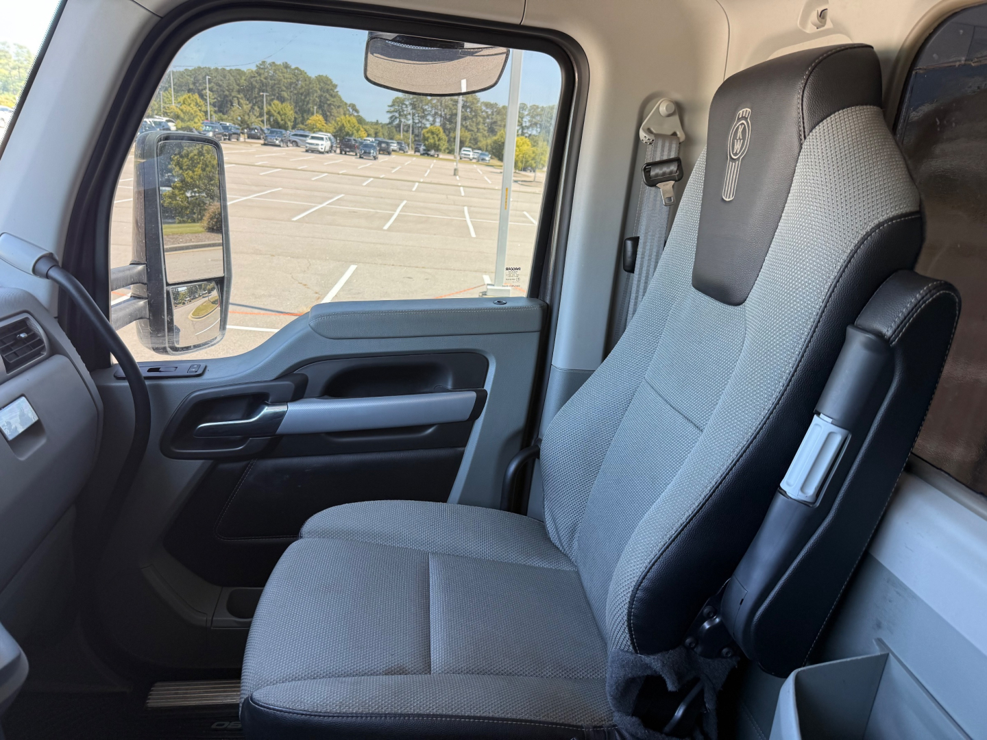 Kenworth T880 truck cabin interior with driver seat and window view; dashboard and side mirror visible; light gray fabric seat with integrated headrest.