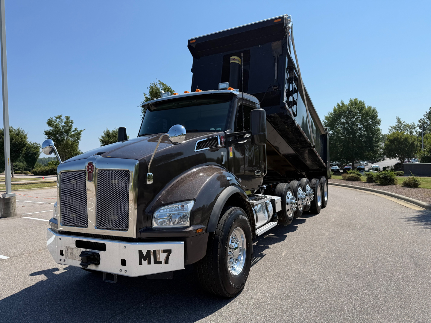 Kenworth T880 dump truck with black cab and raised dump bed, front view in parking lot; ML7 on front bumper, chrome grille, dual axles, chrome mirrors.