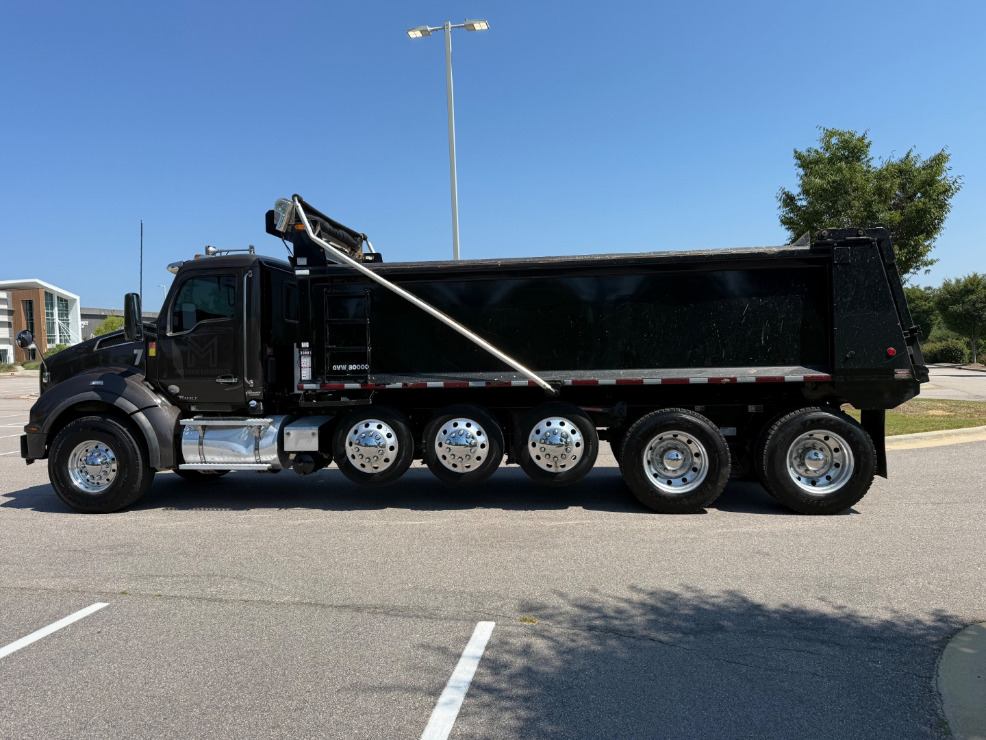 Kenworth T880 dump truck side view showing black dump body, multi-axle rear with six wheels, hydraulic lifting mechanism, chrome fuel tanks, and sleeper-less cab.