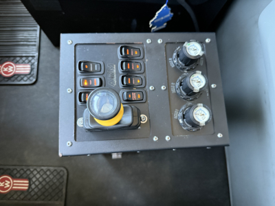 Kenworth T880 control panel showing multiple switches, a yellow-tocked joystick, and three hydraulic gauge knobs.
