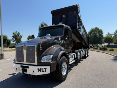 Kenworth T880 dump truck with black cab and raised dump bed, front view in parking lot; ML7 on front bumper, chrome grille, dual axles, chrome mirrors.