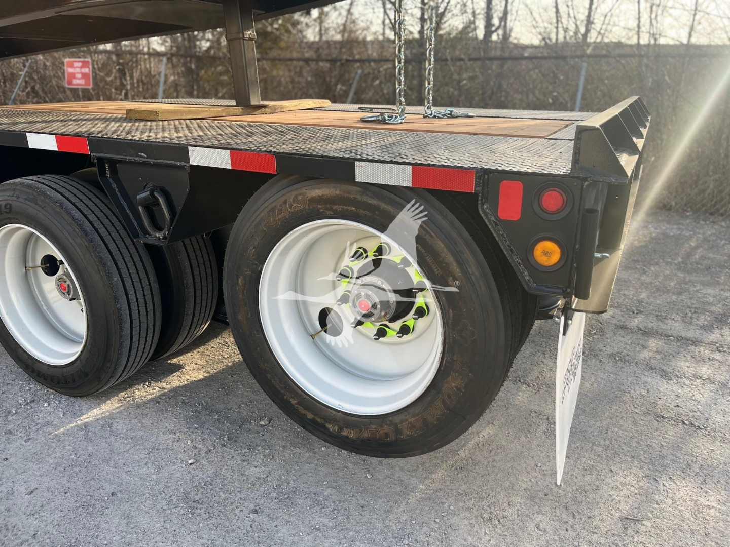 Trailer undercarriage showing two white wheels with black tires, rear fender, taillights, and reflective strip on a lowboy trailer. visible components include wheel hubs and steel deck edge.