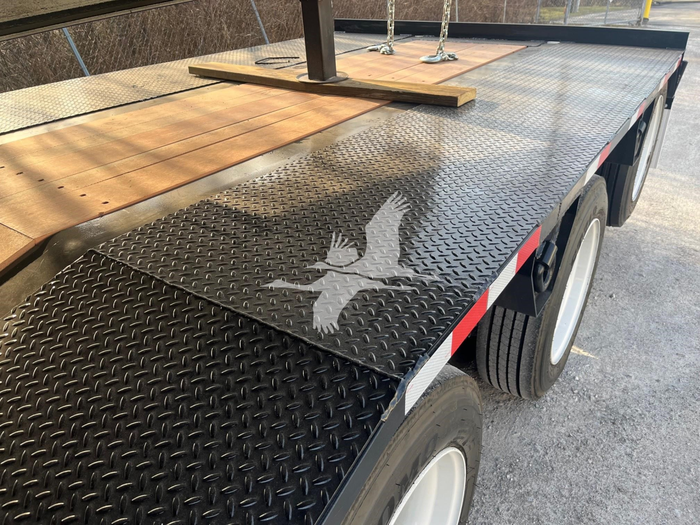 Flatbed trailer with diamond plate steel deck showing a folded wooden ramp and tandem axle wheels.
