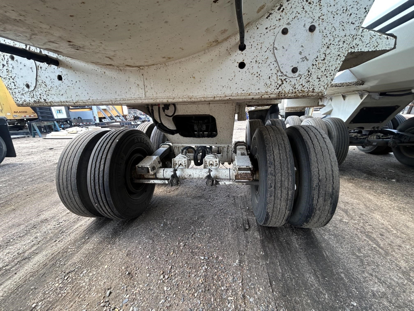 Dump trailer undercarriage showing tandem axle wheels and suspension, with visible leaf springs and metal frame; no text readable on components.