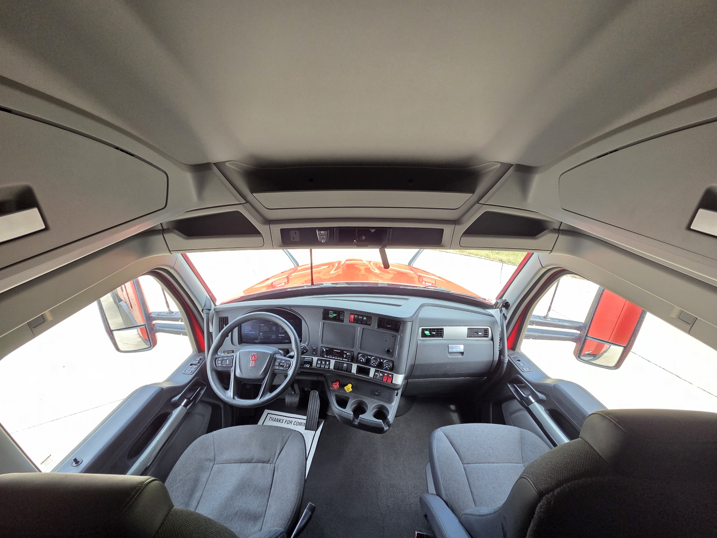 Kenworth T680 cab interior view from driver seat showing steering wheel, dashboard, and front seats. Cabin details visible with sun visors and overhead storage.