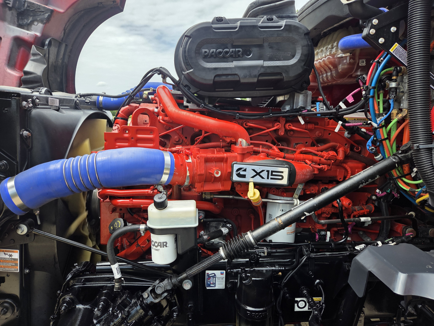 Kenworth T680 engine bay showing red X15 engine, blue intercooler hose, white coolant reservoir, and surrounding wiring.