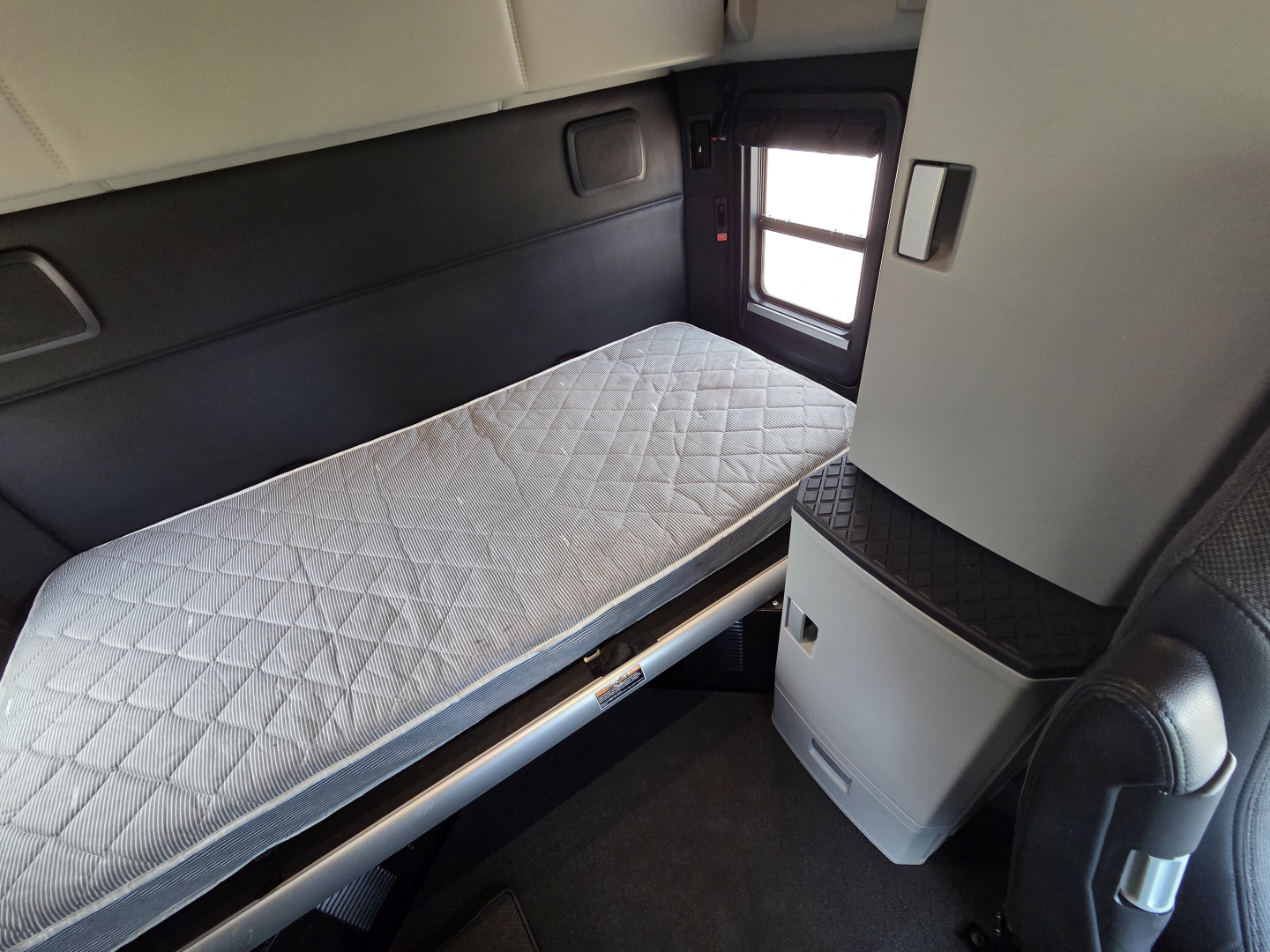 Kenworth T680 sleeper cabin interior with single bunk mattress, small window, and storage cabinet in view.