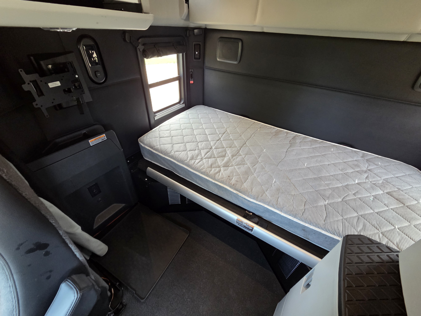 Kenworth T680 sleeper berth cabin interior with single raised bed and window, displaying control panel and compact door area.