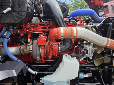 Kenworth T680 engine bay with red engine, blue hoses, and large beige intake tube. Visible components include turbocharger, intercooler piping, alternator, and various hoses.