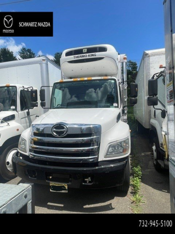Front view of a white 2018 Hino 338 reefer truck with Thermo King unit on roof, chrome grille, black bumper, parked between cargo trucks.