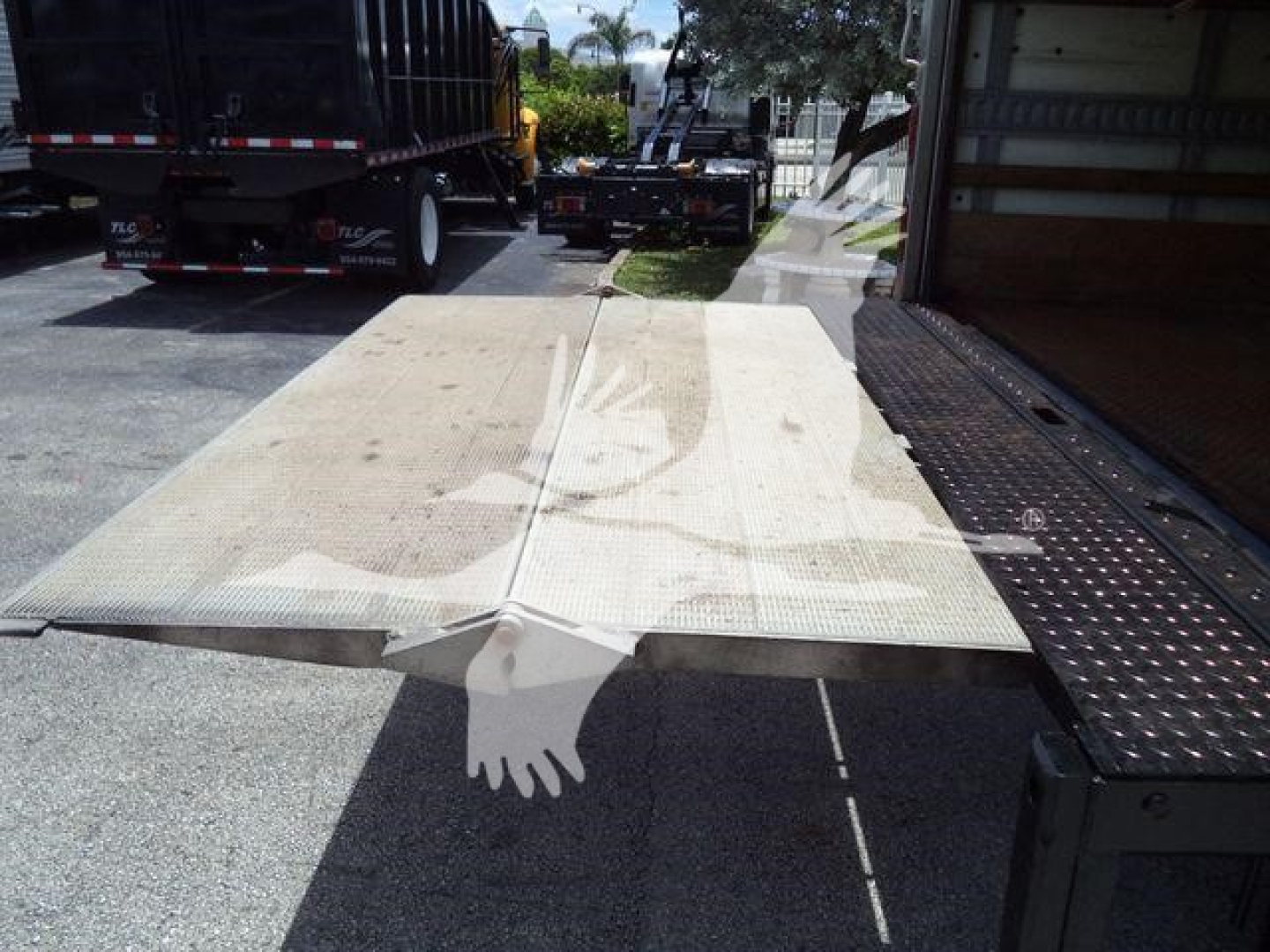 Platform truck bed with loading ramp extended, Isuzu NPR HD context; visible dirt and a hand silhouette watermark overlay in the foreground.
