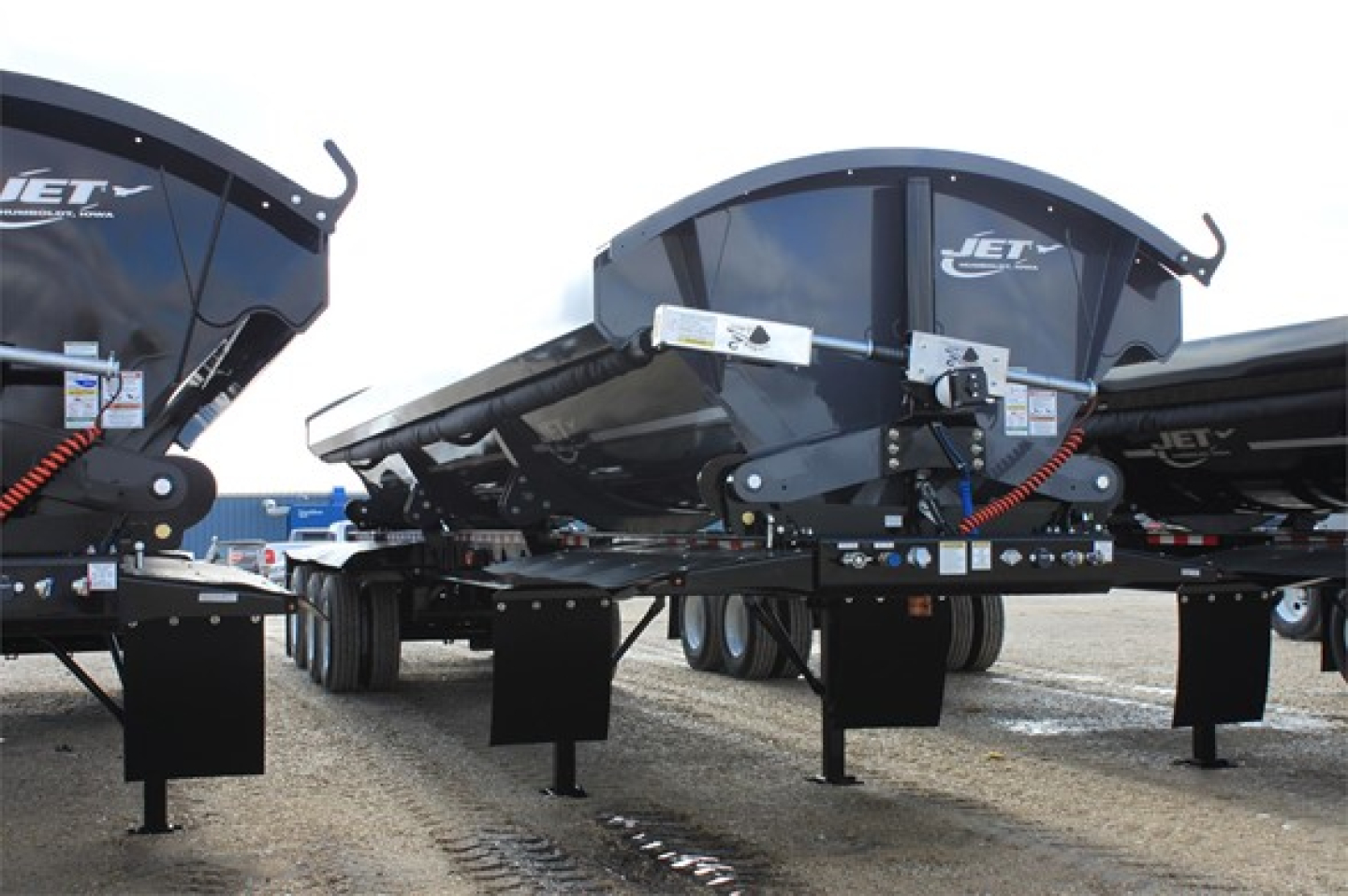Jet tri-axle super spread side dump trailer showing hydraulic components and dual side-mounted dumps.