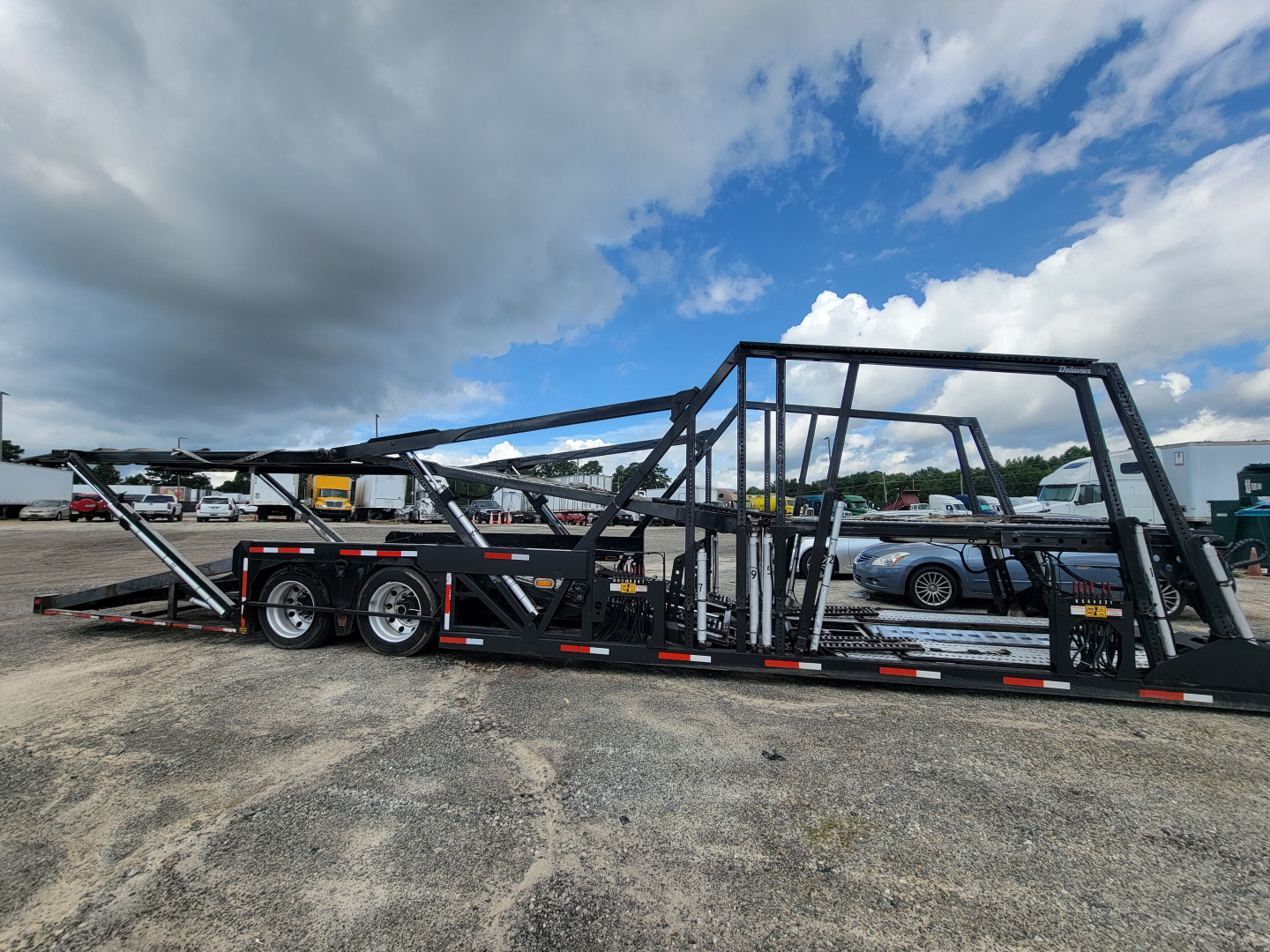 2019 Peterbilt 389 auto carrier trailer in a car-hauler configuration with multiple rails and bunks, parked on a gravel lot under a cloudy sky.