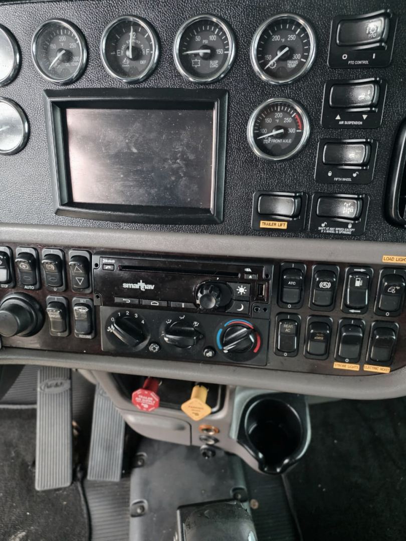 Peterbilt 389 dashboard showing multiple gauges, digital display, climate controls, and switches in the center console; part of the instrument panel with steering column area visible