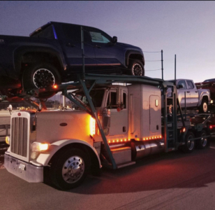 Truck carrier showing a white Peterbilt with a car hauler trailer loaded with a dark blue pickup on top, front grill, side lights, and multiple wheels; dusk lighting.