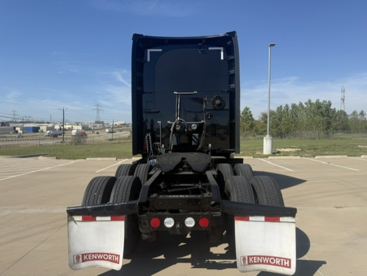 Kenworth T680 rear view showing fifth wheel and dual rear axles with mud flaps and Kenworth branding, back of sleeper cab, clear sky.