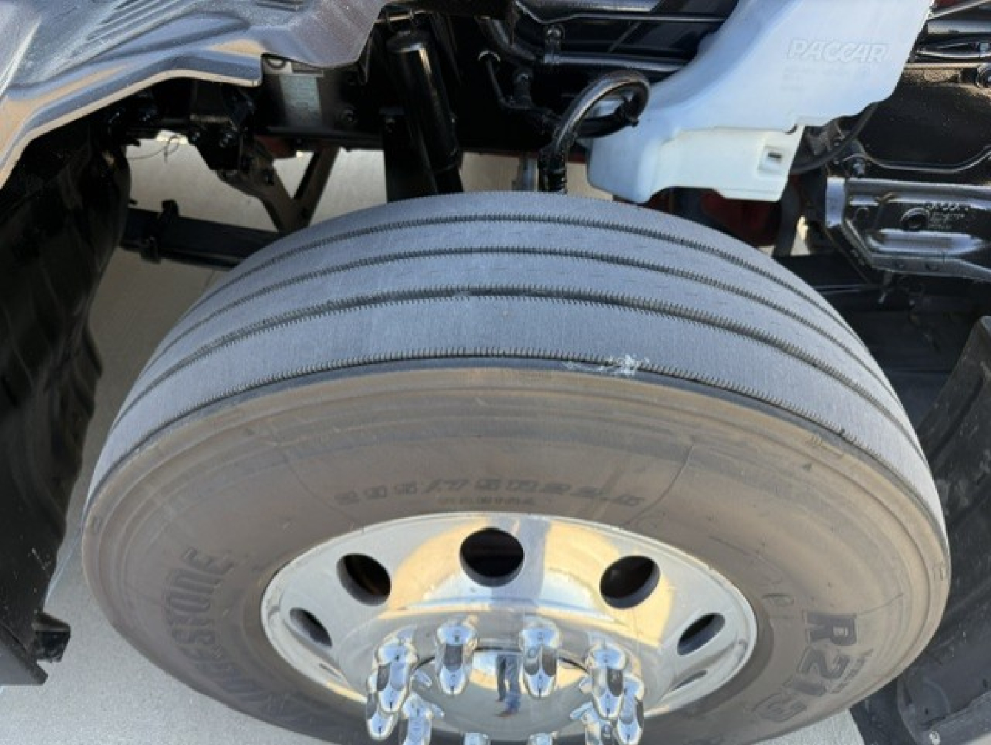 Truck wheel and front axle of a 2023 Kenworth T680 showing dual rear tires and wheel with lug nuts; visible tire sidewall and hub area.