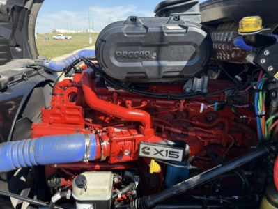 Kenworth T680 engine bay showing Cummins X15 engine with red block and blue intercooler hose, various hoses and components visible.