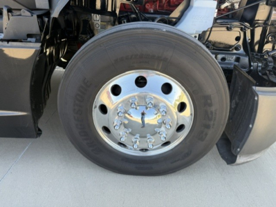 Wheel assembly of a Kenworth T680 truck showing Bridgestone tire and polished chrome hub with lug nuts; front axle area visible with surrounding chassis components.