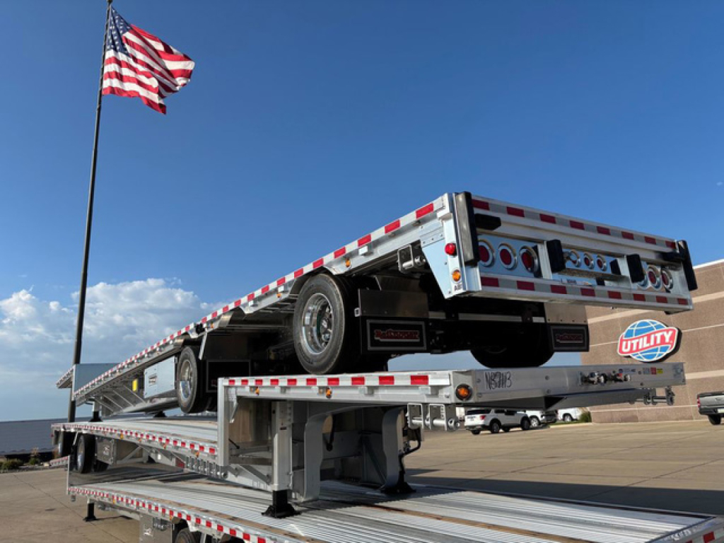 New 2027 Reitnouer dropdeck trailer on a tilt in a yard with an American flag; dual axles, reflective tape, alloy wheels, utility logo on side.