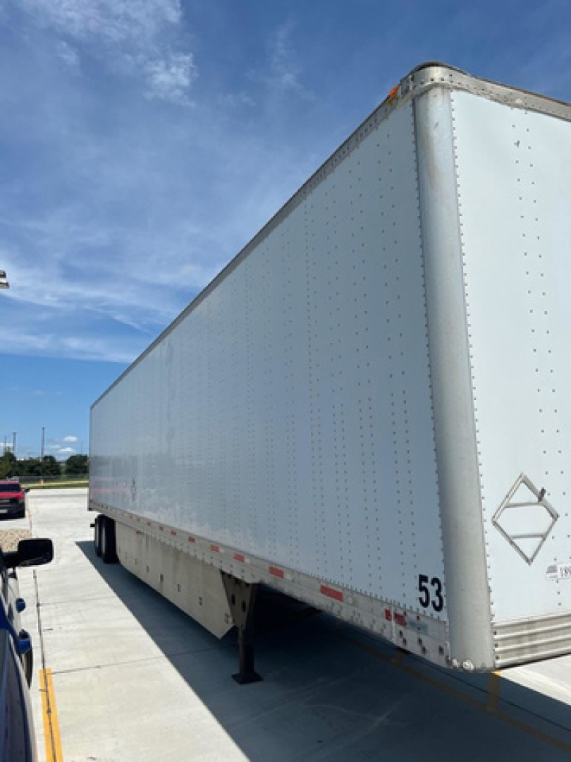Box trailer side view showing white riveted aluminum panel cargo trailer with landing legs and a small diamond hazard sign near rear; number 53 on lower side.