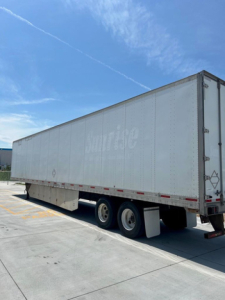Trailer side view showing a large rectangular cargo trailer with dual rear axles and a white exterior; faint markings on side; clear blue sky.