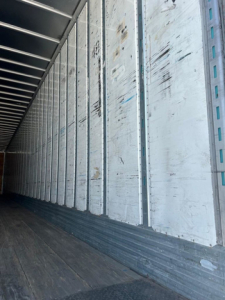 Interior of a used van trailer showing worn metal side walls with vertical ribs and a wooden floor.