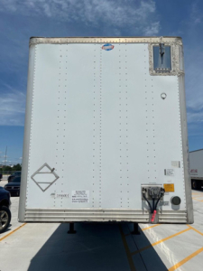 Rear view of a white utility van trailer showing back doors, riveted panels, and a rear bumper with electrical/air connections and reflective stickers.