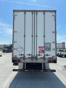 Rear view of a white utility van/trailer showing closed double doors with metal hinges, safety stickers, and rear lights.