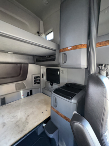 Truck cab interior showing sleeper area with small cabinet, window, and control panel. Visible text includes buttons and labels on the wall and a small screen; no legible VIN or sticker text clearly readable in this view.