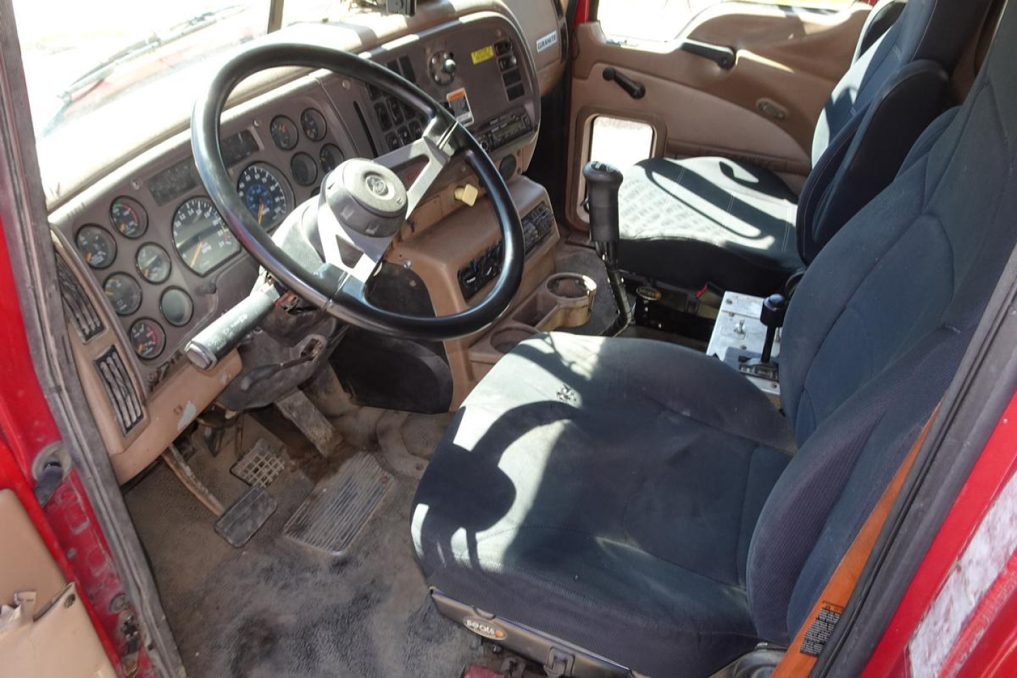 Cabin interior of a Mack CV (Granite) dump truck showing steering wheel, dashboard gauges, and driver seat with gear shifter visible.