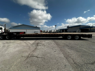 Trailer side view showing a long lowboy drop-deck trailer with triple axle at rear and a flatbed section extending to the horizon.
