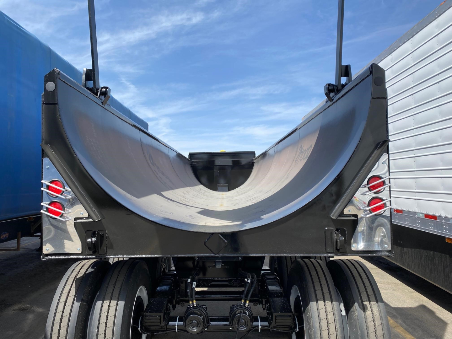 Frameless steel rock end dump trailer bay showing rear end with curved steel body and dual axles, taillights, and hydraulic components.