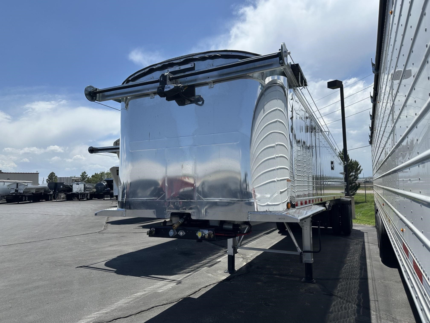 Trailer rear view showing polished aluminum end dump with hydraulic hoist and rear bumper, visible frame and landing gear.