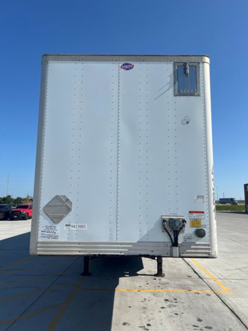 Utility van trailer rear view showing hitch, safety decals, vents, and identification stickers on a white corrugated metal surface.