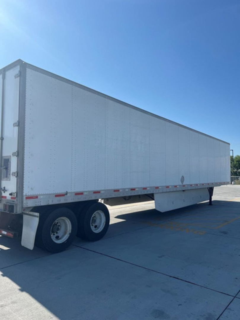 Utility van trailer showing long white box with tandem axles on a concrete lot, side view; no visible text or markings.
