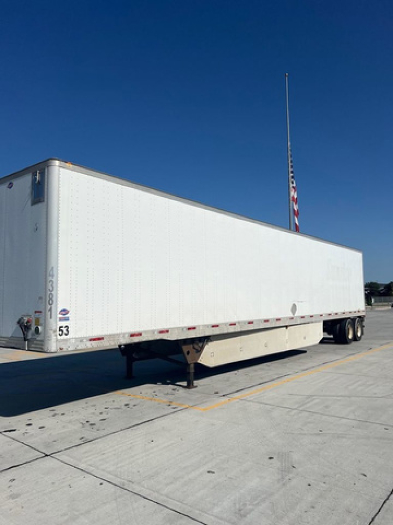 White trailer with long rectangular cargo area, support legs down, black tires at rear, small numbers near front corner, and a tall flagpole in background under a clear blue sky.