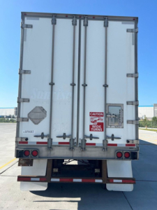 Rear view of a white utility van trailer showing double rear doors, external latches, taillights, and safety decals; visible CAUTION/WIDE TURNS sticker and license-like markings.