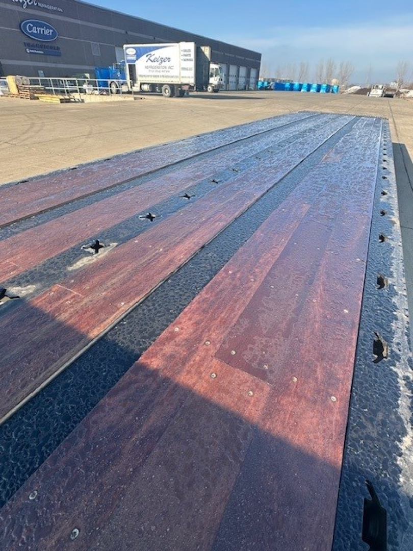Long flatbed trailer bed with wooden planks and metal edge, viewed from the front, with a Carrier building and trucks in the background.