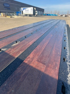 Long flatbed trailer bed with wooden planks and metal edge, viewed from the front, with a Carrier building and trucks in the background.