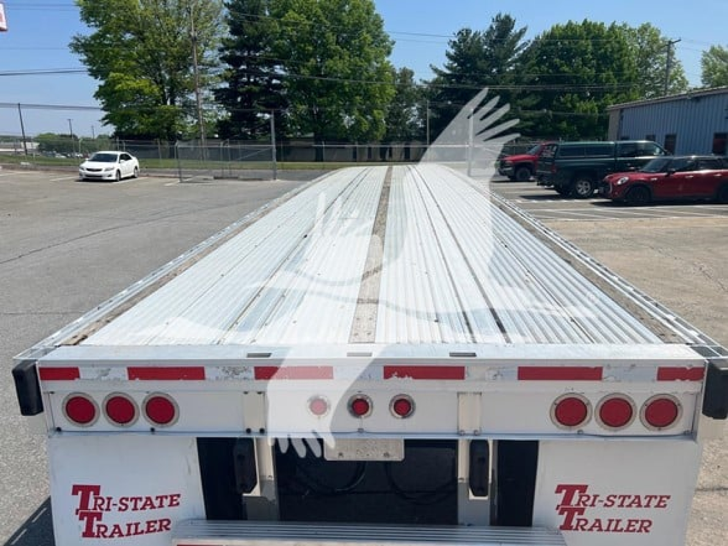 Flatbed trailer back view showing metal planks and rear lights, Tri-State Trailer branding on left, stock plate, and long bed with reflective markings.