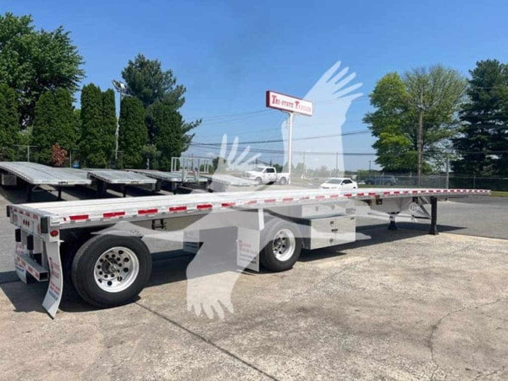 Flatbed trailer with long extendable deck and dual rear axles, white with red reflective tape, parked on lot under trees.