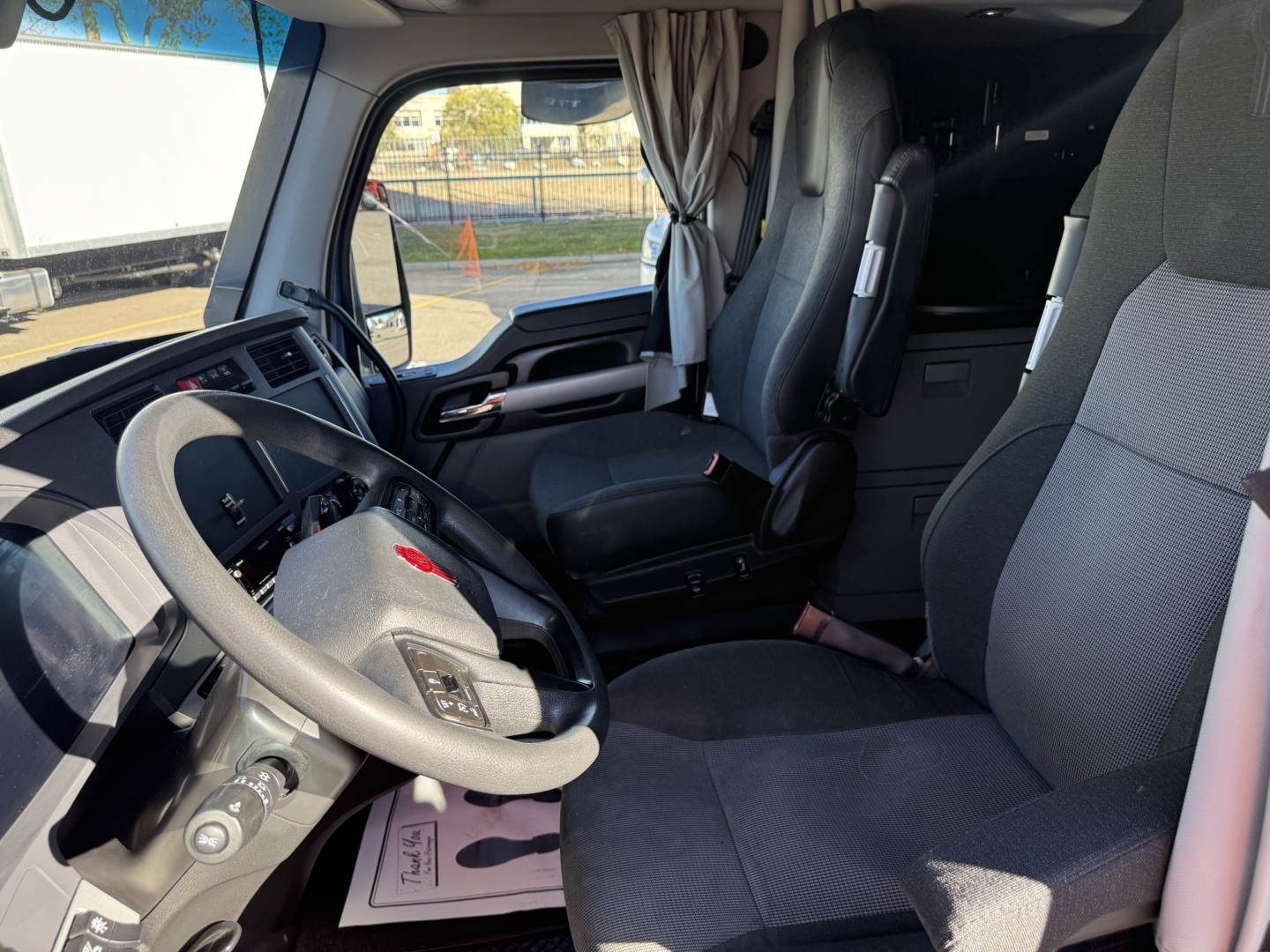 Truck cab interior view with steering wheel, dashboard, and seats; curtain behind; no visible labels or numbers readable.