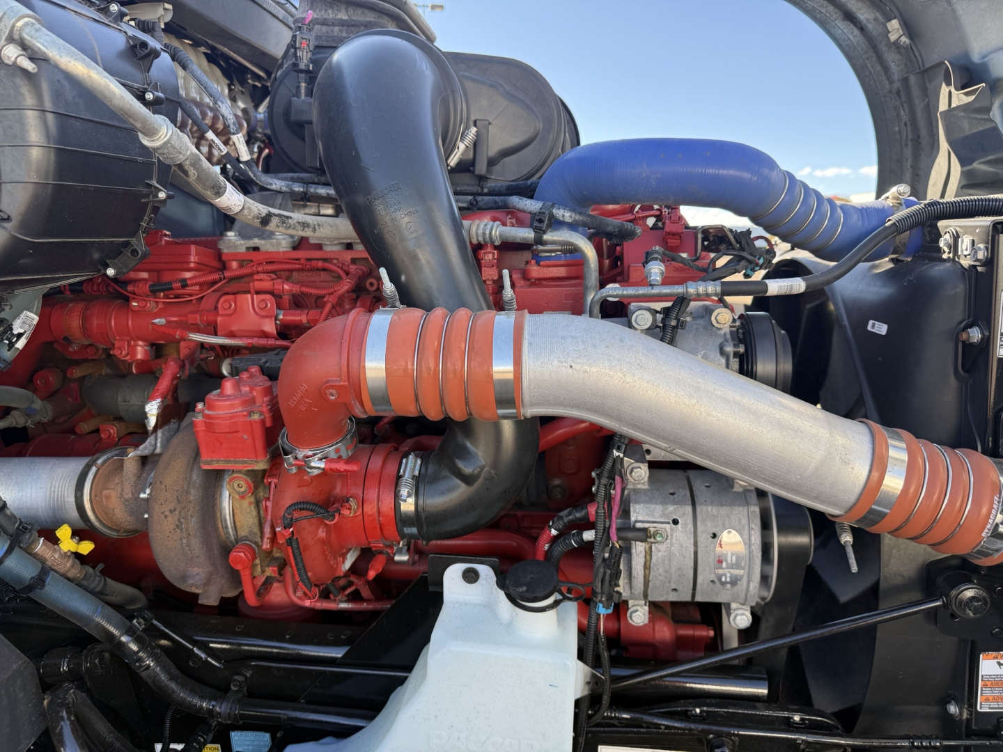 Kenworth T680 engine bay showing red engine components, large silver exhaust duct, blue hoses, and black intake piping.
