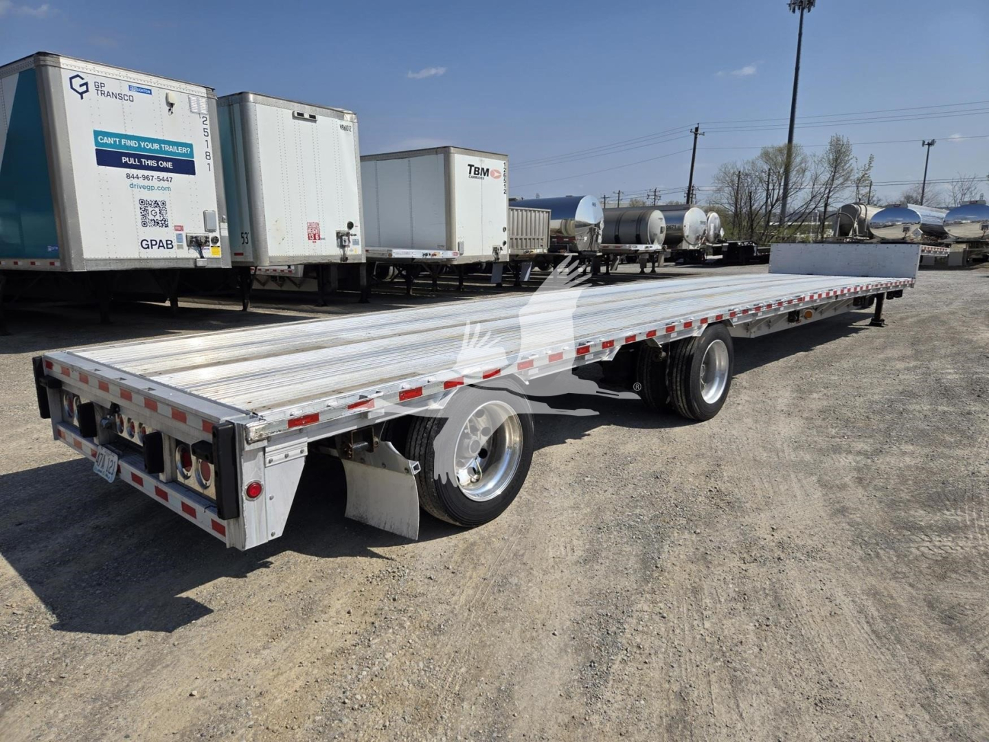 Flatbed trailer in a dealer lot with drop deck, dual axles, aluminum sides, and rear lighting. Various equipment trailers and boxes visible in background.